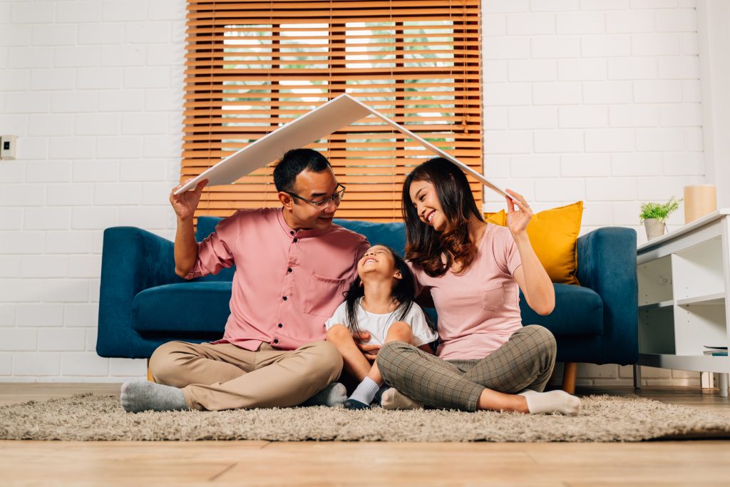Happy family holding symbolic cardboard roof of life insurance and sitting on couch at home, Family care, Protection and safety life insurance concept, Mortgage loan and investment security for future, home warranty