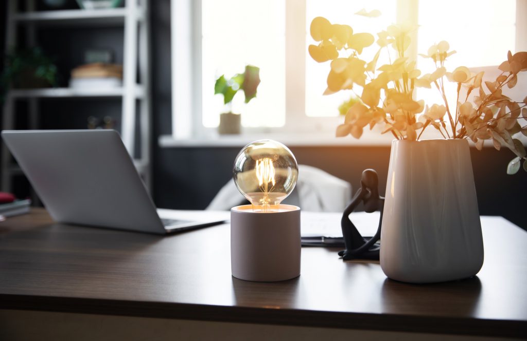 A cozy workspace with a laptop on a wooden desk, a glowing light bulb, and a vase of dried flowers. Sunlight softly filters through the window, creating a warm and inviting atmosphere. energy efficient windows